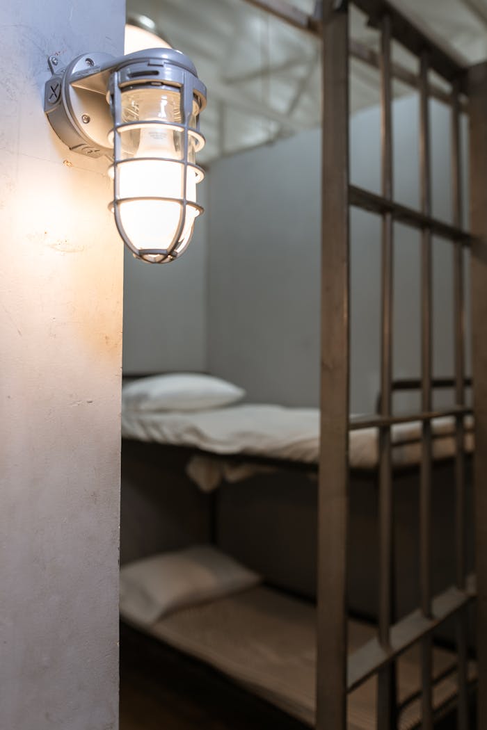 Home Interior of a jail cell featuring a steel bunk bed and an industrial wall lamp.
